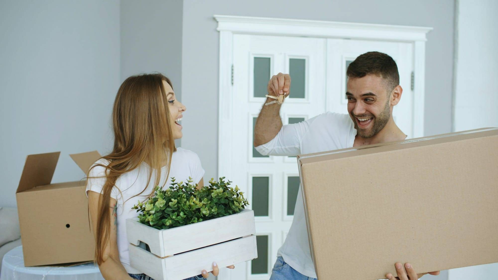 Un couple rit au milieu des cartons : lui brandit un trousseau de clés, elle porte une caisse de plantes.